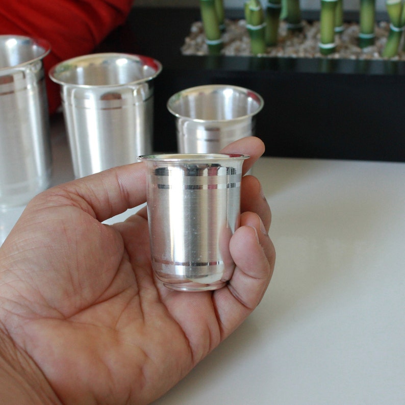 May include: Four silver drinking glasses with a decorative band around the middle. The glasses are different sizes and are on a white surface.