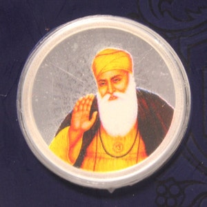 May include: A silver coin with a portrait of a man with a white beard and a yellow turban. The man is wearing a yellow robe and has a gold necklace with a pendant.