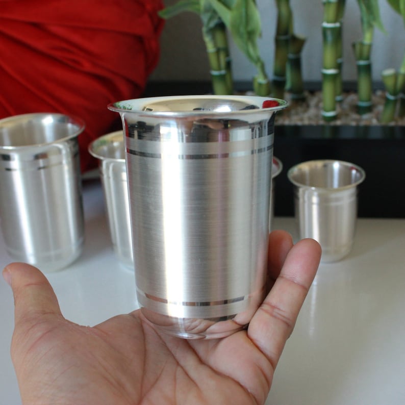 May include: A set of four silver drinking glasses with a brushed finish and a silver band around the top. The glasses are on a white surface.