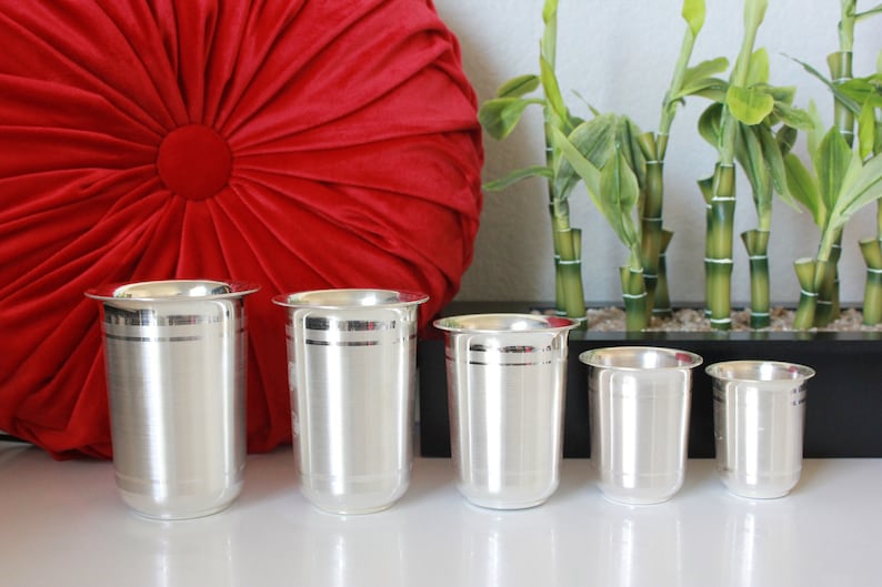 May include: Five silver drinking glasses of varying sizes, with a decorative band around the middle of each glass. The glasses are arranged in a row on a white surface.