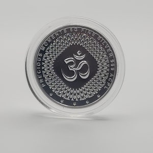 May include: A silver coin with a circular design featuring the Om symbol in the center. The coin is encased in a clear plastic capsule. The text "PRECIOUS MOMENTS 5G FINE SILVER 999 AC 21" is inscribed around the edge of the coin.