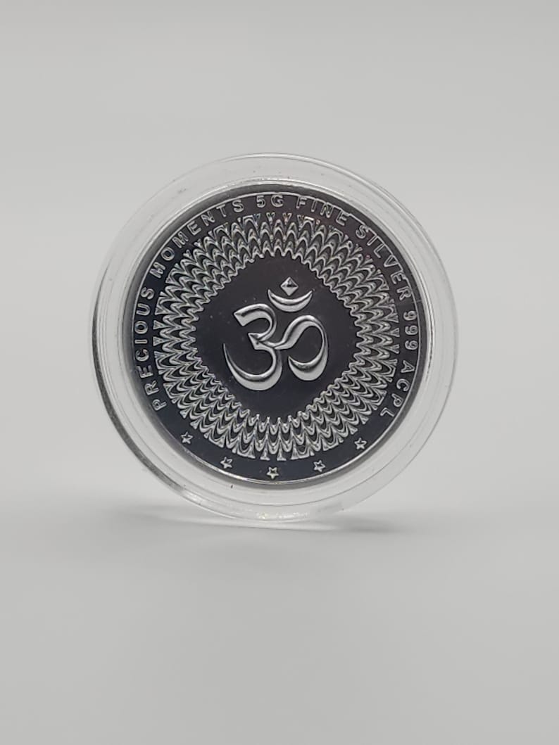 May include: A silver coin with a circular design featuring the Om symbol in the center. The coin is encased in a clear plastic capsule. The text "PRECIOUS MOMENTS 5G FINE SILVER 999 AC 21" is inscribed around the edge of the coin.
