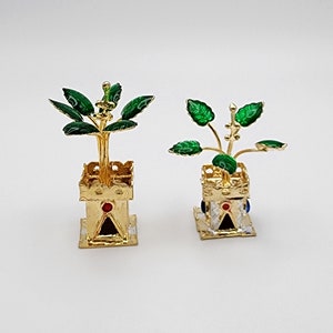 May include: Two gold-colored metal figurines of potted plants. The plants have green leaves and are in gold-colored pots with red and blue accents.