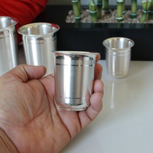 May include: Four silver drinking glasses with a decorative band around the middle. The glasses are on a white surface.