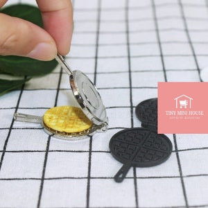 May include: A miniature waffle iron with a silver finish and a black waffle iron with a waffle pattern. The waffle iron is open and a waffle is inside. The waffle iron is on a white and black checkered surface.