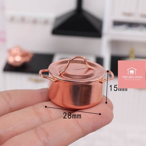 Könnte beinhalten: Ein Miniatur-Kupfertopf mit Deckel und Griff. Der Topf ist 28 mm breit und 15 mm hoch.  "TINY MINI HOUSE" ist auf einem rosa Quadrat im Hintergrund gedruckt.
