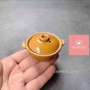May include: A small, brown ceramic pot with a lid. The pot has a small hole in the lid. The pot is being held in a hand.