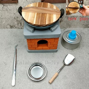 Op de afbeelding: Miniatuur keukenset met een wok, fornuis en accessoires. De wok heeft een houten deksel en staat op een grijs en oranje bakstenen fornuis. Extra items zijn een kleine metalen kom, spatel, pincet en een brandstofpatroon.