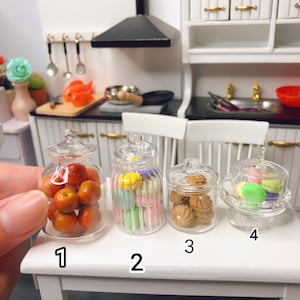Könnte beinhalten: Vier durchsichtige Glasbehälter mit Deckeln, gefüllt mit bunten Süßigkeiten. Die Behälter sind mit 1, 2, 3 und 4 beschriftet. Die Behälter stehen auf einer weißen Oberfläche.