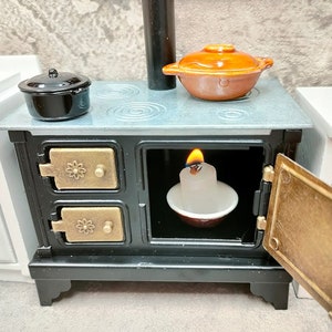 Puede incluir: Una estufa en miniatura negra con una parte superior gris, dos puertas de latón y una vela blanca en el interior. La estufa tiene una olla en la parte superior y una cacerola con tapa en el lado derecho.
