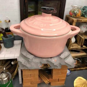 May include: A pink ceramic pot with a black lid sits on a miniature brick stove. The stove has a gray stone top and is made of red bricks.