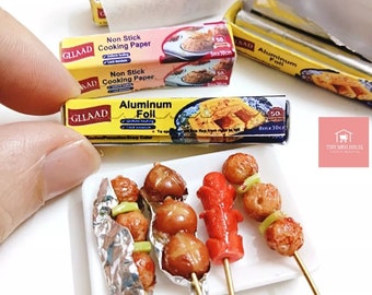 May include: A miniature set of three skewers with grilled meat and vegetables, wrapped in aluminum foil. The skewers are on a white plate. There are three boxes of miniature cooking supplies in the background, labeled "GLLAAD Non Stick Cooking Paper" and "GLLAAD Aluminum Foil".