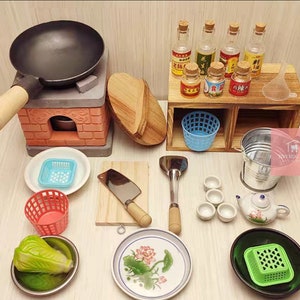 Op de afbeelding: Miniatuur keukenset met een zwarte wok op een bakstenen fornuis, houten snijplank, hakmes en diverse kleine schalen. Inclusief een kleine theepot, kommen en flessen met etiketten. Een charmante collectie voor poppenhuizen of knutselwerk.