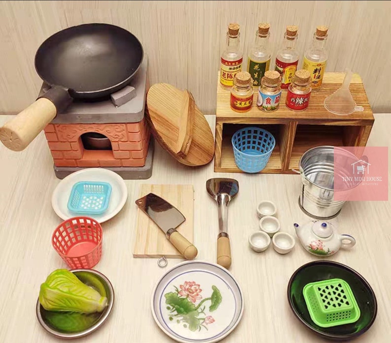 Op de afbeelding: Miniatuur keukenset met een zwarte wok op een bakstenen fornuis, houten snijplank, hakmes en diverse kleine schalen. Inclusief een kleine theepot, kommen en flessen met etiketten. Een charmante collectie voor poppenhuizen of knutselwerk.