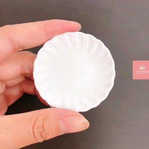 May include: A small, white ceramic plate with a fluted edge. The plate is about 2 inches in diameter.