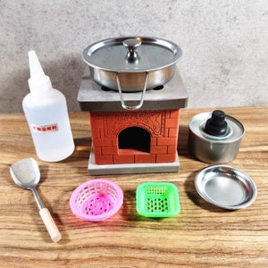Op de afbeelding: Een miniatuur kookset met een kleine roestvrijstalen pan met deksel, een rode bakstenen kachel en diverse accessoires. Inclusief een fles met tekst, een spatel en kleine roze en groene mandjes. De set is ontworpen om mee te spelen of te etaleren.