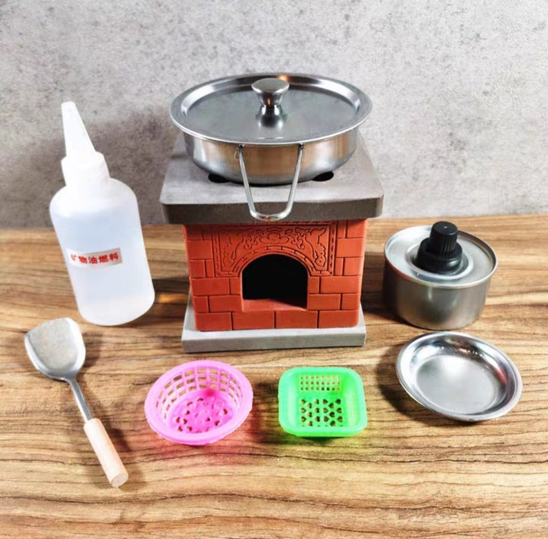 Op de afbeelding: Een miniatuur kookset met een kleine roestvrijstalen pan met deksel, een rode bakstenen kachel en diverse accessoires. Inclusief een fles met tekst, een spatel en kleine roze en groene mandjes. De set is ontworpen om mee te spelen of te etaleren.