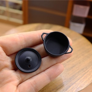 Puede incluir: Un juego de olla y tapa en miniatura de color negro para cocinas de casas de muñecas. La olla tiene dos asas y una forma redonda. La tapa es plana y redonda.