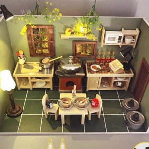 Op de afbeelding: Miniatuurkeukendiorama met een groen en wit betegelde vloer. De sc&egrave;ne omvat een fornuis, magnetron, tafelsetting en diverse keukengerei. De muren zijn groen geschilderd en de sc&egrave;ne wordt verlicht door kleine lampen.