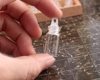 May include: A clear glass vial with a white plastic stopper and a small loop on top. The vial is small and is being held in a hand.
