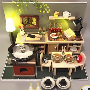Op de afbeelding: Miniatuurkeuken met een fornuis, tafel en planken. Het fornuis heeft een pot met een deksel en een houten handvat. De tafel is gedekt voor twee personen met kommen en lepels. Er zijn kleine flesjes en een lamp.
