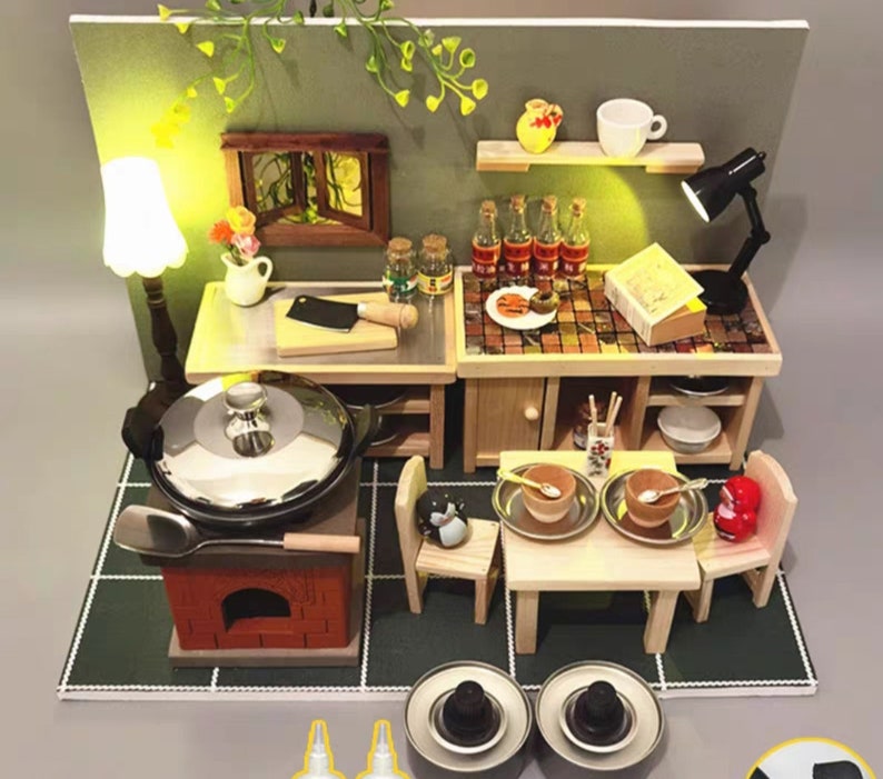 Op de afbeelding: Miniatuurkeuken met een fornuis, tafel en planken. Het fornuis heeft een pot met een deksel en een houten handvat. De tafel is gedekt voor twee personen met kommen en lepels. Er zijn kleine flesjes en een lamp.