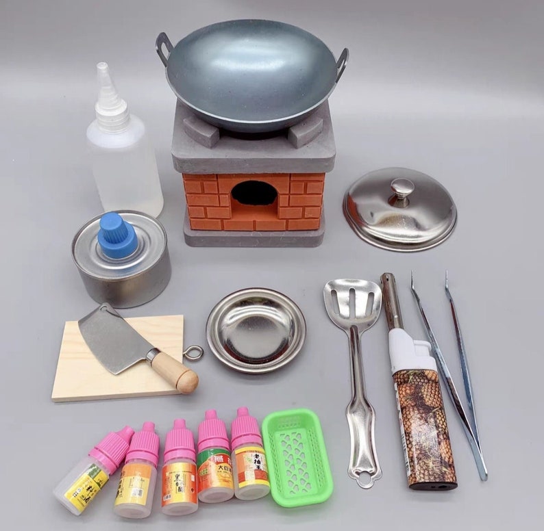 Op de afbeelding: Een miniatuurkeuken speelset met een grijze wok, een bakstenen fornuis, een spatel, een klein metalen bord, een pincet, een aansteker, een klein metalen deksel, een blikje brandstof, een klein houten snijplank met een zilveren mes, een klein groen mandje en vijf kleine flesjes vloeistof.