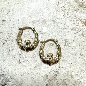 May include: A pair of gold hoop earrings with a Celtic knot design. The knots are in the shape of clasped hands.