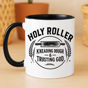 Peut inclure: Mug en céramique blanche avec une anse et un bord noirs. Le mug présente le texte "HOLY ROLLER" au-dessus d'une illustration d'un rouleau à pâtisserie, avec les mots "KNEADING DOUGH & TRUSTING GOD" en dessous.