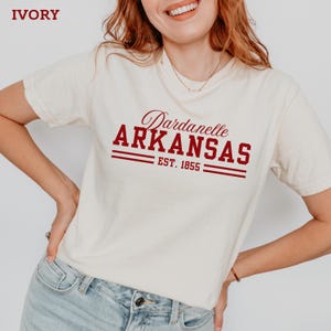 May include: Ivory-colored t-shirt with the text "Dardanelle ARKANSAS EST. 1855" in burgundy. The shirt has short sleeves and a crew neck. The text is in a serif font.