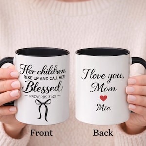 Könnte beinhalten: Zwei weiße Keramikbecher mit schwarzen Griffen und Rändern. Auf einem Becher steht "Her children rise up and call her Blessed" mit einer Schleifengrafik. Der andere Becher trägt die Aufschrift "I love you, Mom" mit einem roten Herzen und dem Namen "Mia".