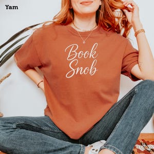 May include: A woman wearing a rust-colored t-shirt with the text "Book Snob" in white script. She is wearing blue jeans and has her hair pulled back.