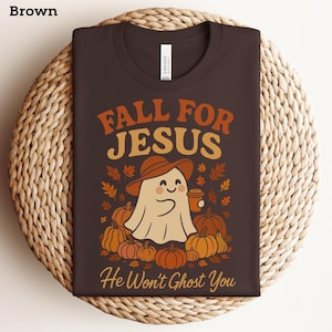 May include: Brown t-shirt featuring the text "FALL FOR JESUS" in orange and yellow. A cartoon ghost wearing a hat and holding a coffee cup is surrounded by pumpkins and autumn leaves. The text "He Won't Ghost You" is below.