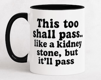 May include: White ceramic mug with a black interior and handle. The mug features the text "This too shall pass.. like a kidney stone, but it'll pass" in bold, black lettering. A humorous and motivational item.