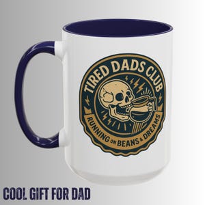 以下が含まれることがあります： ネイビーブルーの内部とハンドルが付いた白いセラミックマグ。マグには、コーヒーカップから飲んでいる骸骨の金とネイビーブルーのグラフィックがあり、「TIRED DADS CLUB」と「RUNNING ON BEANS & DREAMS」の文字が書かれています。「COOL GIFT FOR DAD」の文字が一番下にあります。
