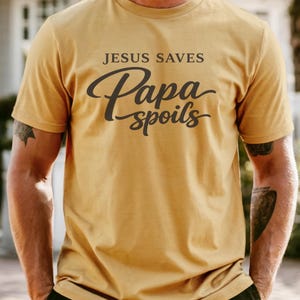 May include: Mustard-colored t-shirt with the text "JESUS SAVES" above "Papa spoils" in a script font. The shirt is short-sleeved and appears to be made of a soft material.