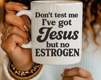 May include: White ceramic mug with the text "Don't test me I've got Jesus but no ESTROGEN" in black. The mug has a curved handle and is held by a person.