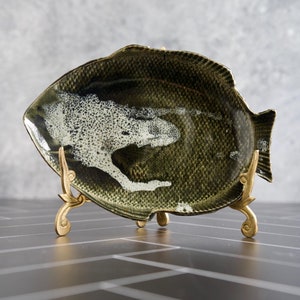 May include: A green and white ceramic fish-shaped serving dish with a textured surface, displayed on a gold metal stand.
