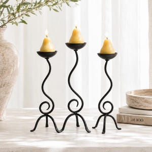 May include: Three black metal candle holders with curved, swirling stems and tripod bases. Each holder features a round cup supporting a pale yellow, pear-shaped candle. The candles are lit, casting a soft glow from the top of each holder.