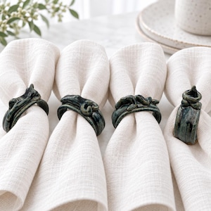 May include: Four off-white linen napkins folded and held by individual dark green ceramic napkin rings. Each ring features a unique, hand-sculpted, organic, and textured design with a glossy finish, showcasing variations in the deep green glaze.