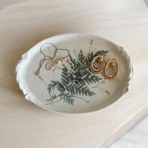 May include: A white ceramic oval tray with a fern design and gold jewelry. The tray has a gold necklace, a pearl necklace, and two gold hoop earrings. The fern design is in shades of green and brown.