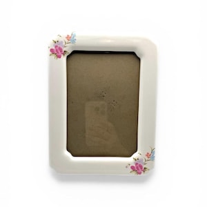 May include: A rectangular white ceramic picture frame with a floral design in the corners. The flowers are pink, purple, and blue. The frame has a brown interior.