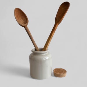 May include: A small, light gray ceramic jar with two wooden spoons inside and a cork lid beside it. The spoons are different shapes and shades of brown. The jar has a rounded body and a narrow neck.