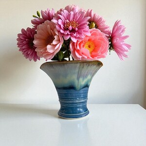 May include: A ceramic vase in shades of blue and green holds a bouquet of pink flowers. The vase has a flared top and a textured, ribbed design. The flowers include dahlias and peonies, with varying shades of pink and coral.