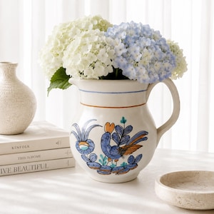 May include: A ceramic pitcher with a cream-colored base, featuring a hand-painted blue and orange bird motif, filled with white and light blue hydrangea flowers. The pitcher has a curved handle and a thin blue and orange horizontal stripe design.