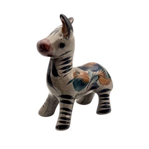 May include: A ceramic zebra figurine with a gray base color and black stripes. The zebra has a dark red nose and is decorated with floral patterns in shades of blue, orange, and green. The figurine is standing on four legs.