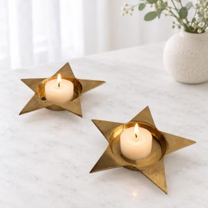 May include: Two star-shaped candle holders made of brass, each featuring a central circular depression holding a single white tea light candle with a lit flame. The metallic finish of the star-shaped holders reflects light, creating a warm glow.