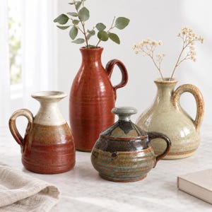 May include: A collection of four ceramic vessels including two tall pitchers with handles, one smaller pitcher with a two-tone cream and rust glaze, and a squat lidded jar with a dark brown and green mottled finish, holding sprigs of greenery.