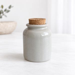 May include: A cylindrical ceramic jar with a smooth, light gray glossy finish and a flat, textured natural cork stopper. The container features a short, slightly flared rim at the top and a flat base, presenting a simple, uniform, and solid structure.