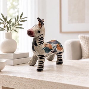 May include: A ceramic zebra figurine featuring a cream-colored base with hand-painted black stripes, a dark red nose, and floral motifs in shades of orange, blue, and green on its back. The four-legged animal sculpture has pointed ears.